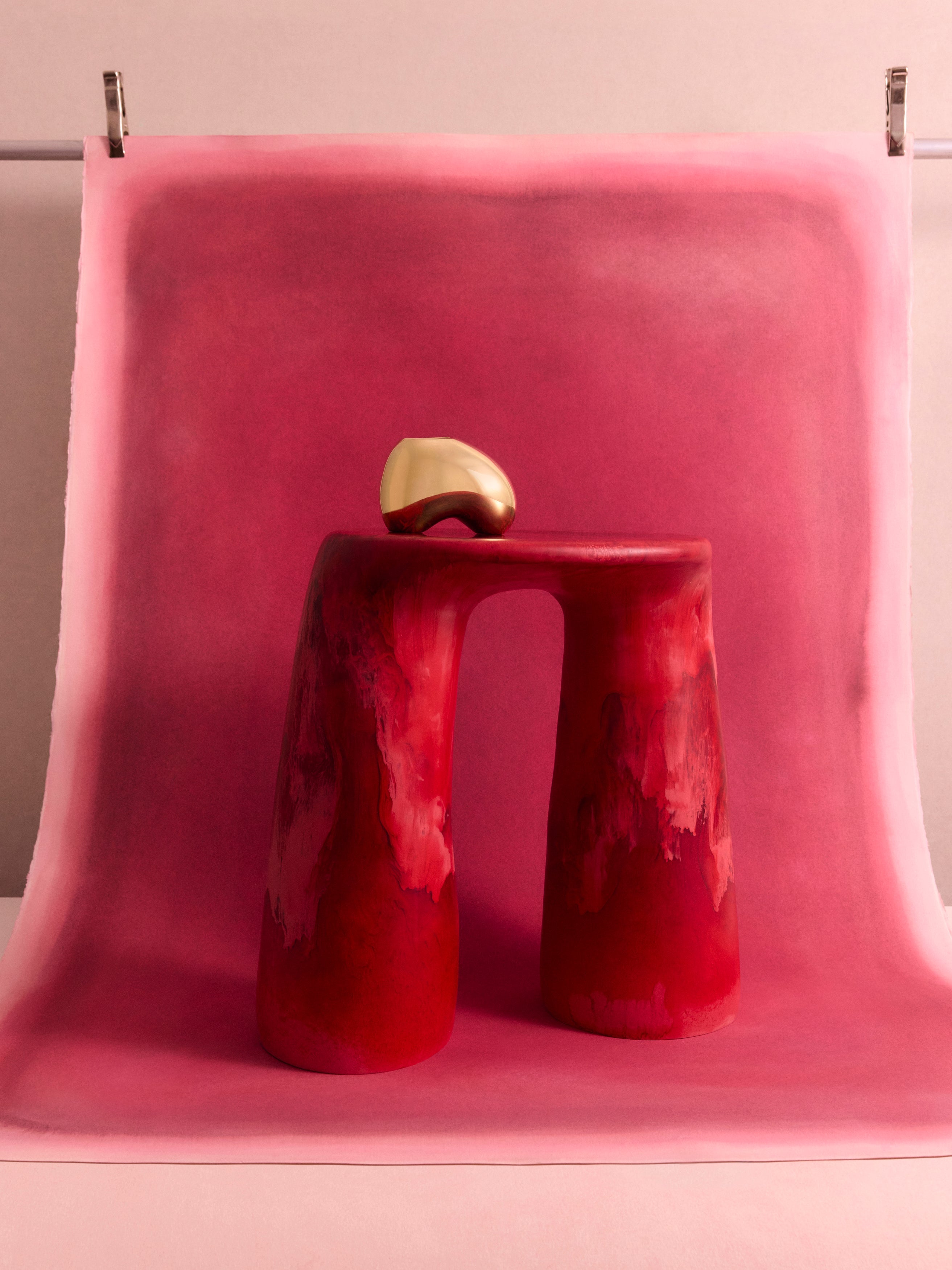 Deep pink two legged resin table with a brass sculptural vase atop on a pink backdrop