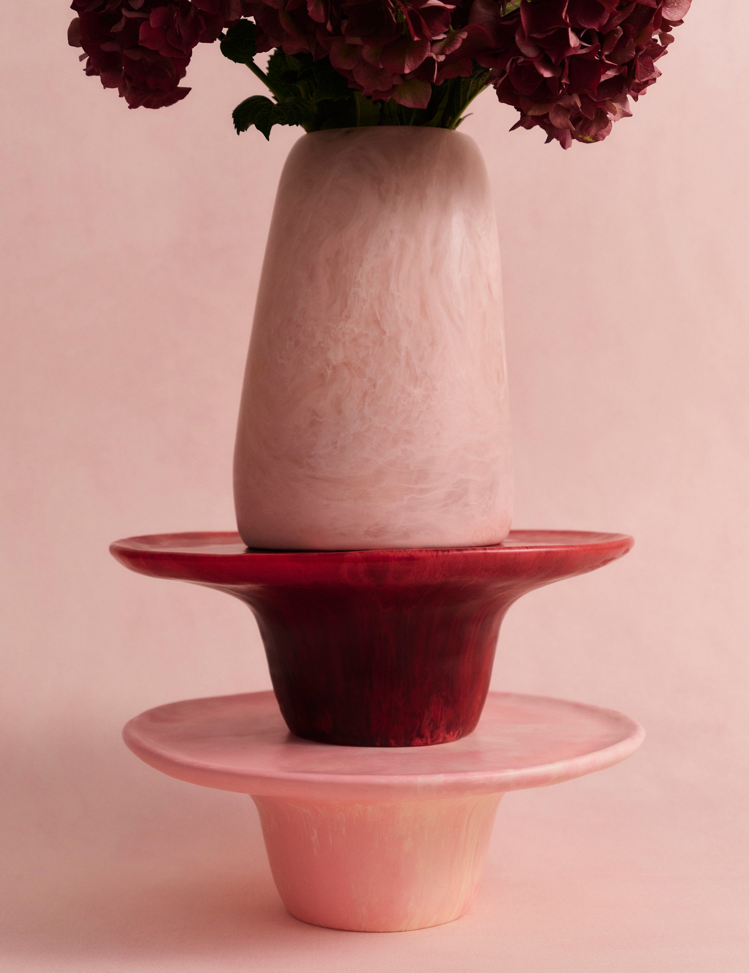 Two pink cake stands with a large pink resin vase atop with deep pink flowers on a light pink backdrop