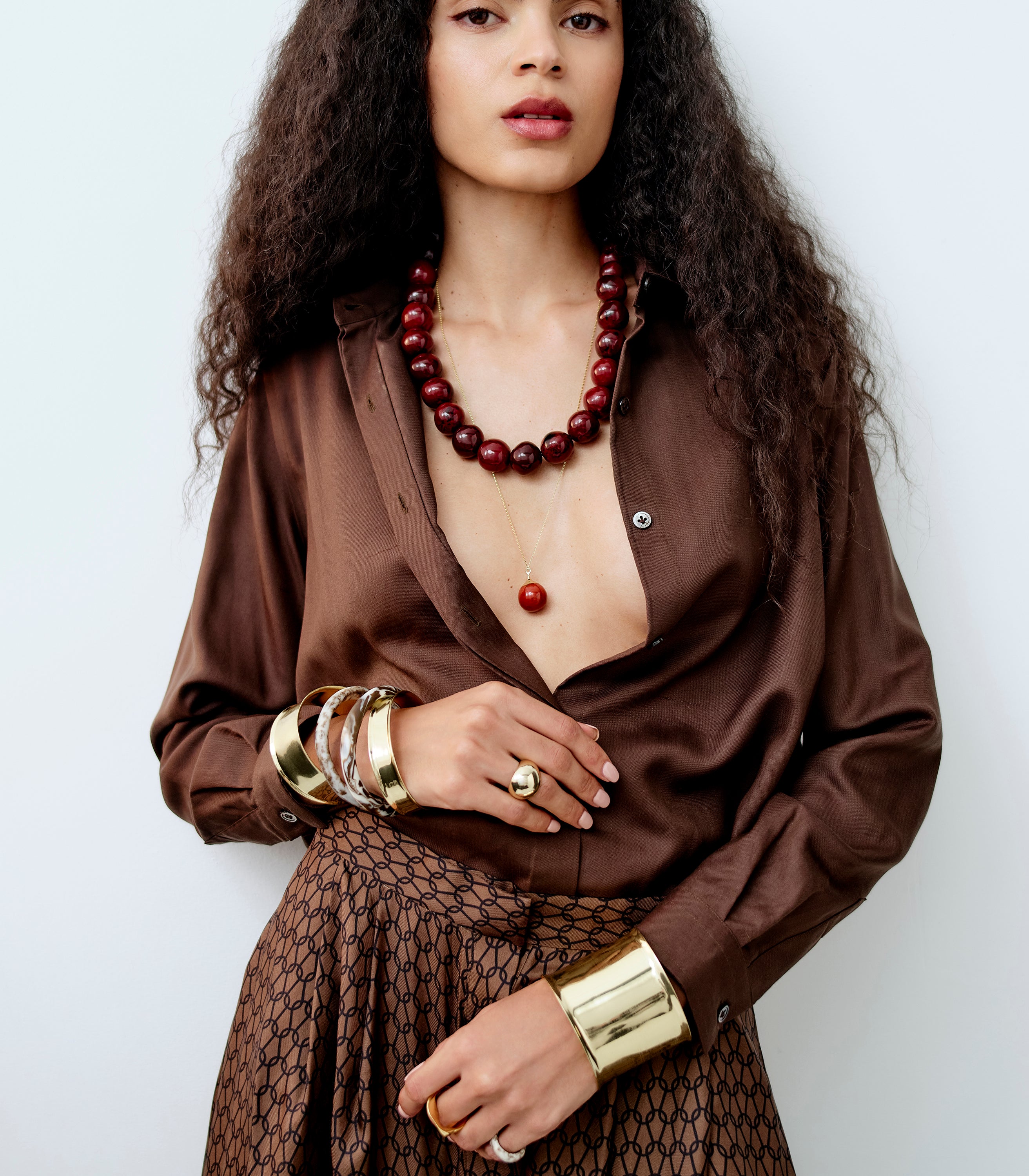 Woman wearing a brown satin blouse with a deep neckline, gold bracelets, and a red beaded necklace against a white background.