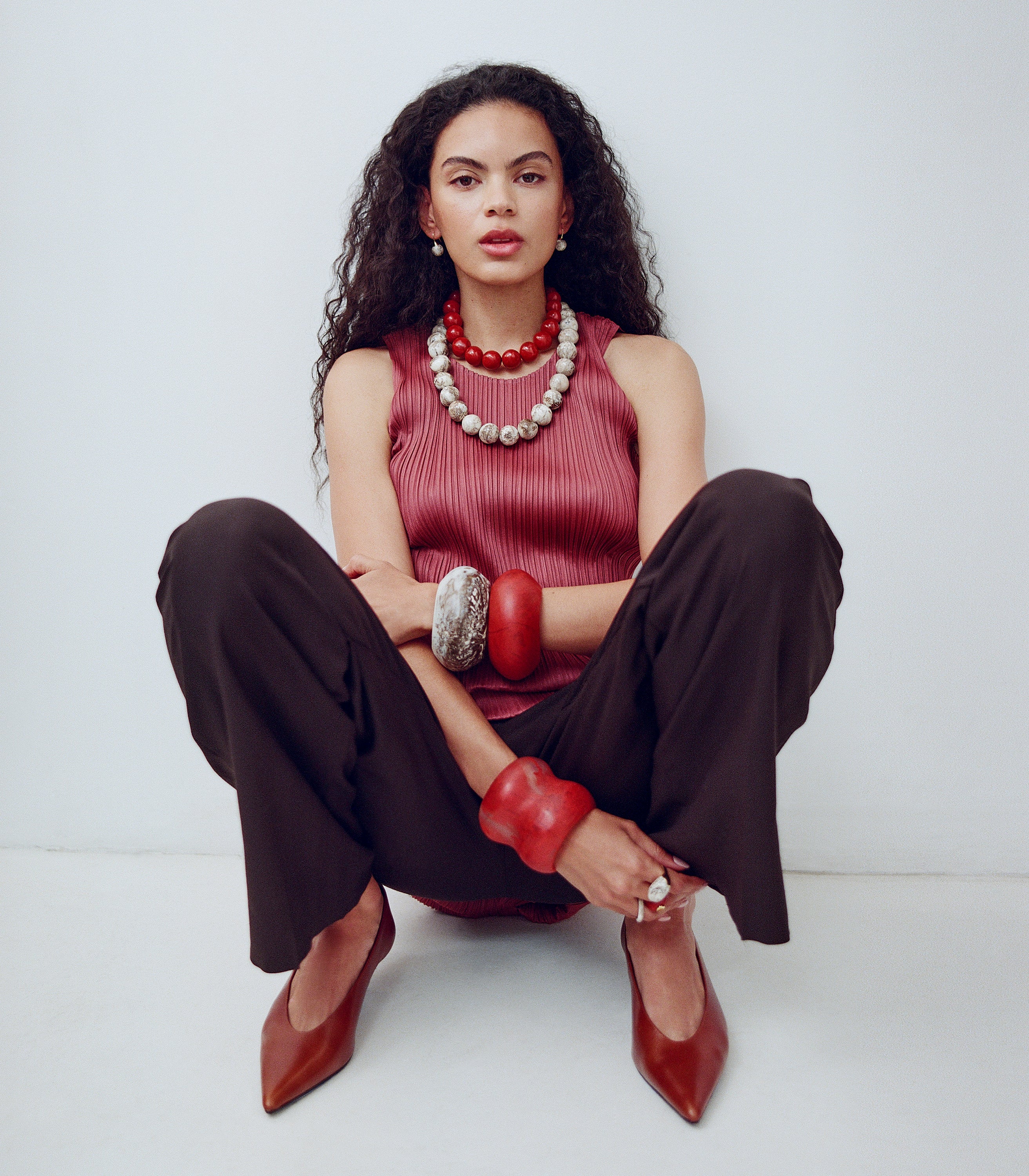 Woman sitting on the floor wearing a red sleeveless top, brown pants, and red shoes.